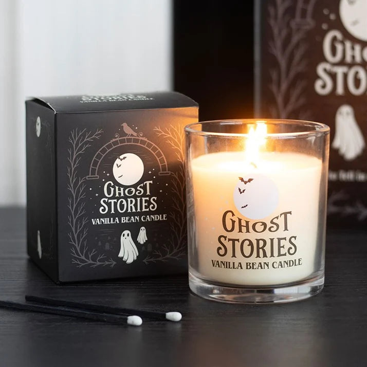 A lit 'Ghost Stories' vanilla bean candle in a clear glass jar sits next to its black packaging box on a dark tabletop. Two cotton swabs are placed in the foreground, with a blurred version of the box in the background.