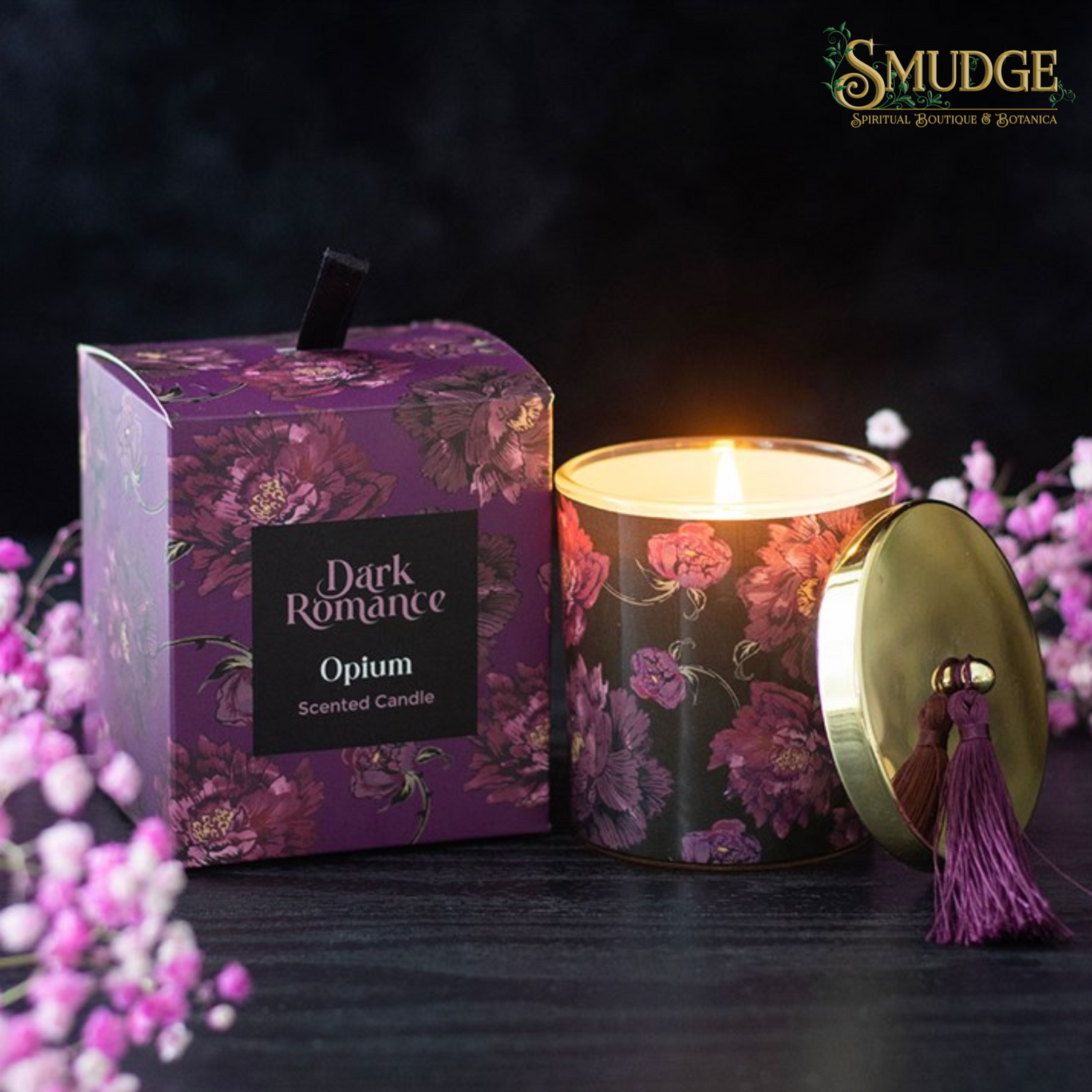 A lit scented candle in a floral-patterned jar beside its matching purple box, with the gold lid placed next to it on a dark surface. The candle and packaging are branded Dark Romance and Smudge.