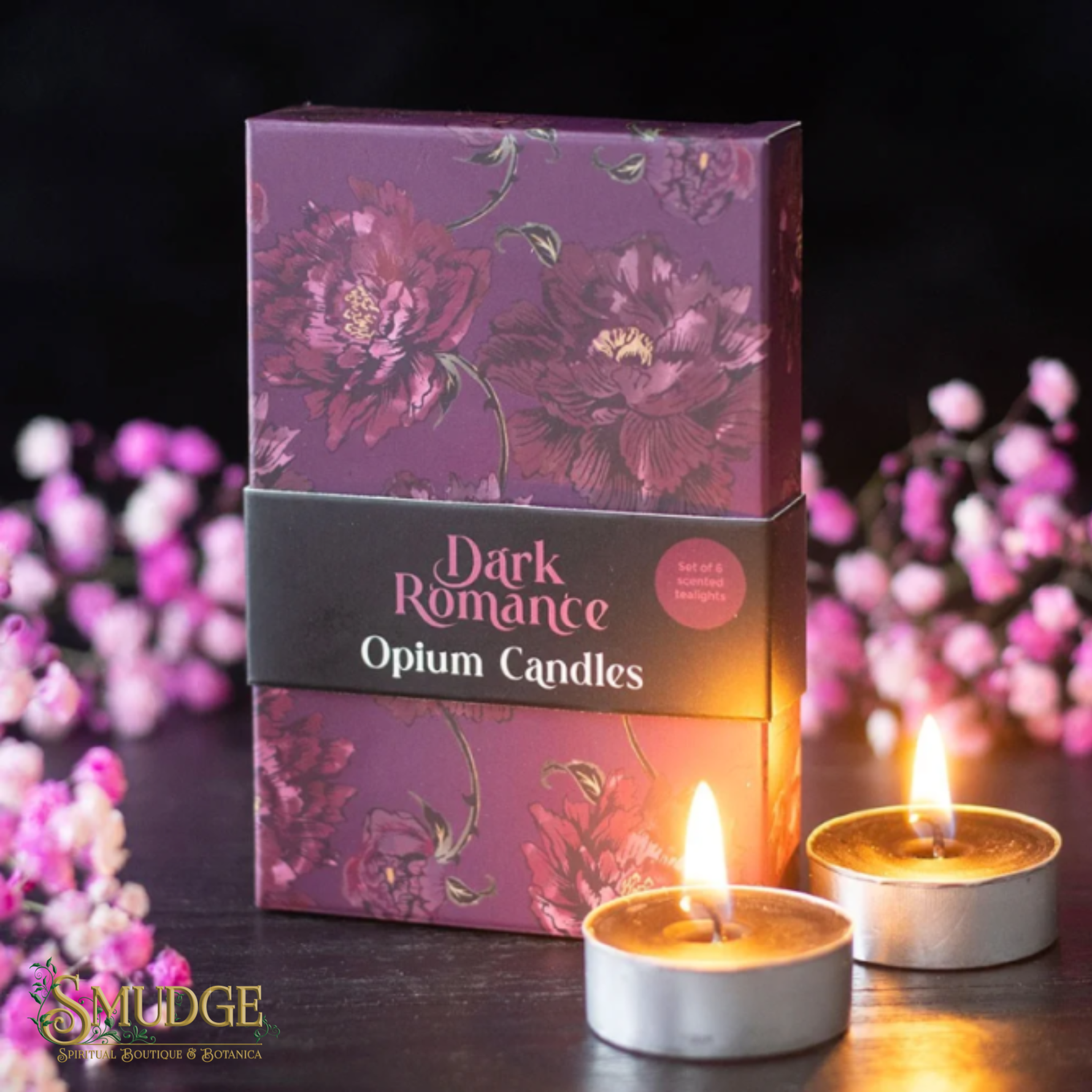 A box of Dark Romance Opium Candles by Smudge is shown on a dark wooden surface with two lit tealight candles in front and pink flowers blurred in the background.
