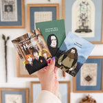 Hand holding four cards with illustrated characters and text in front of a wall with framed pictures.
