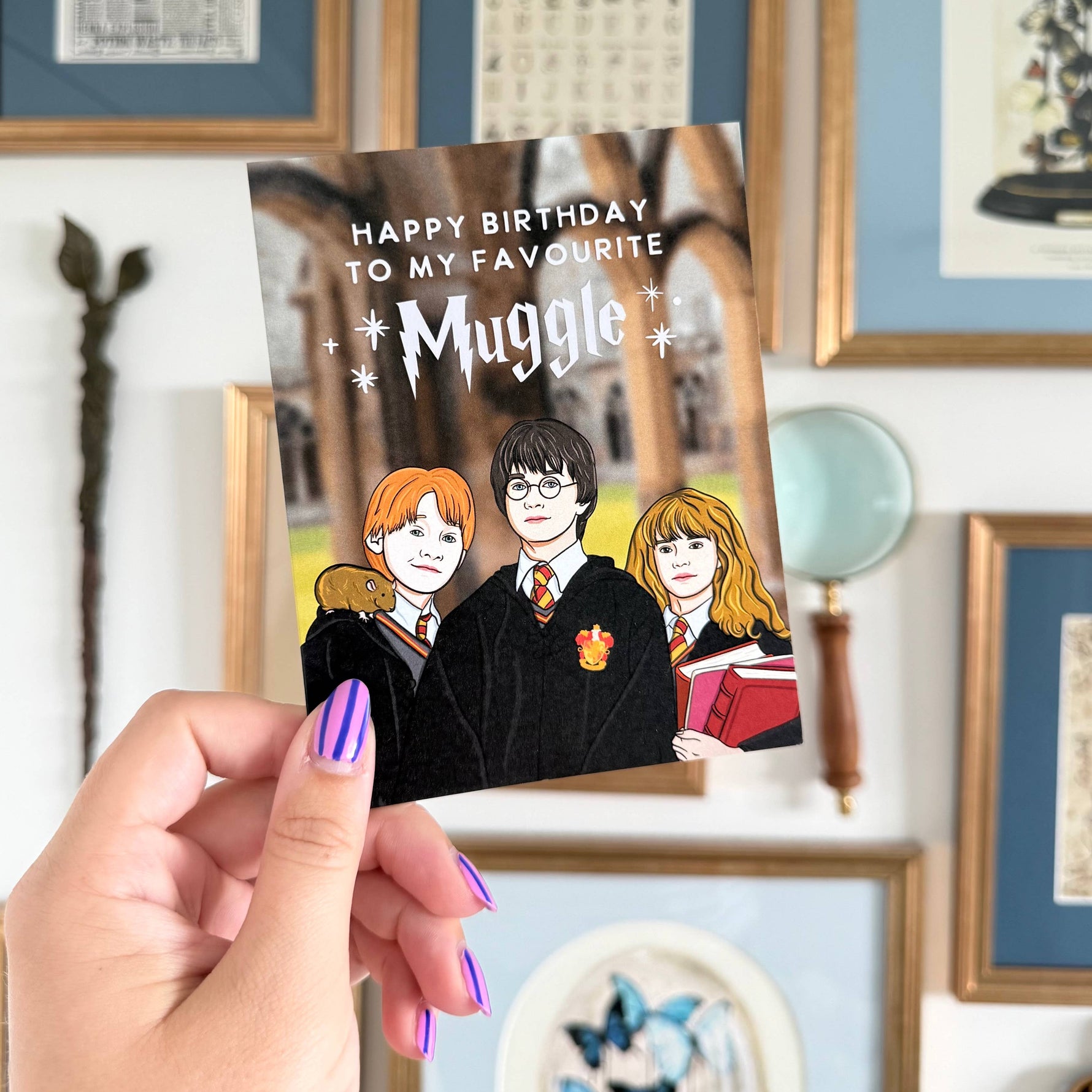Hand holding a card with cartoon characters and 'Happy Birthday to my favourite Muggle' text against a decorative wall.