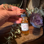 A hand holds a small glass bottle labeled 'Dragon's Blood' by Smudge Spiritual Boutique & Botanica, surrounded by eucalyptus leaves, a purple crystal cluster, and a wooden tray.