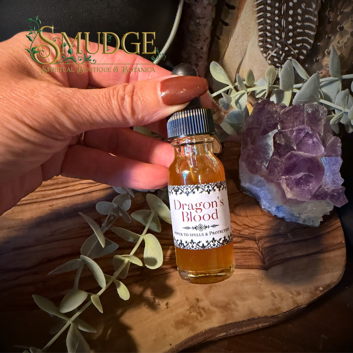 A hand holds a small glass bottle labeled 'Dragon's Blood' by Smudge Spiritual Boutique & Botanica, surrounded by eucalyptus leaves, a purple crystal cluster, and a wooden tray.