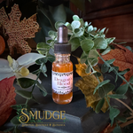A small glass dropper bottle labeled 'Dragon's Blood' by Smudge Spiritual Boutique & Botanica, filled with amber liquid, styled among green leaves, dried leaves, and a white orchid on a dark surface.