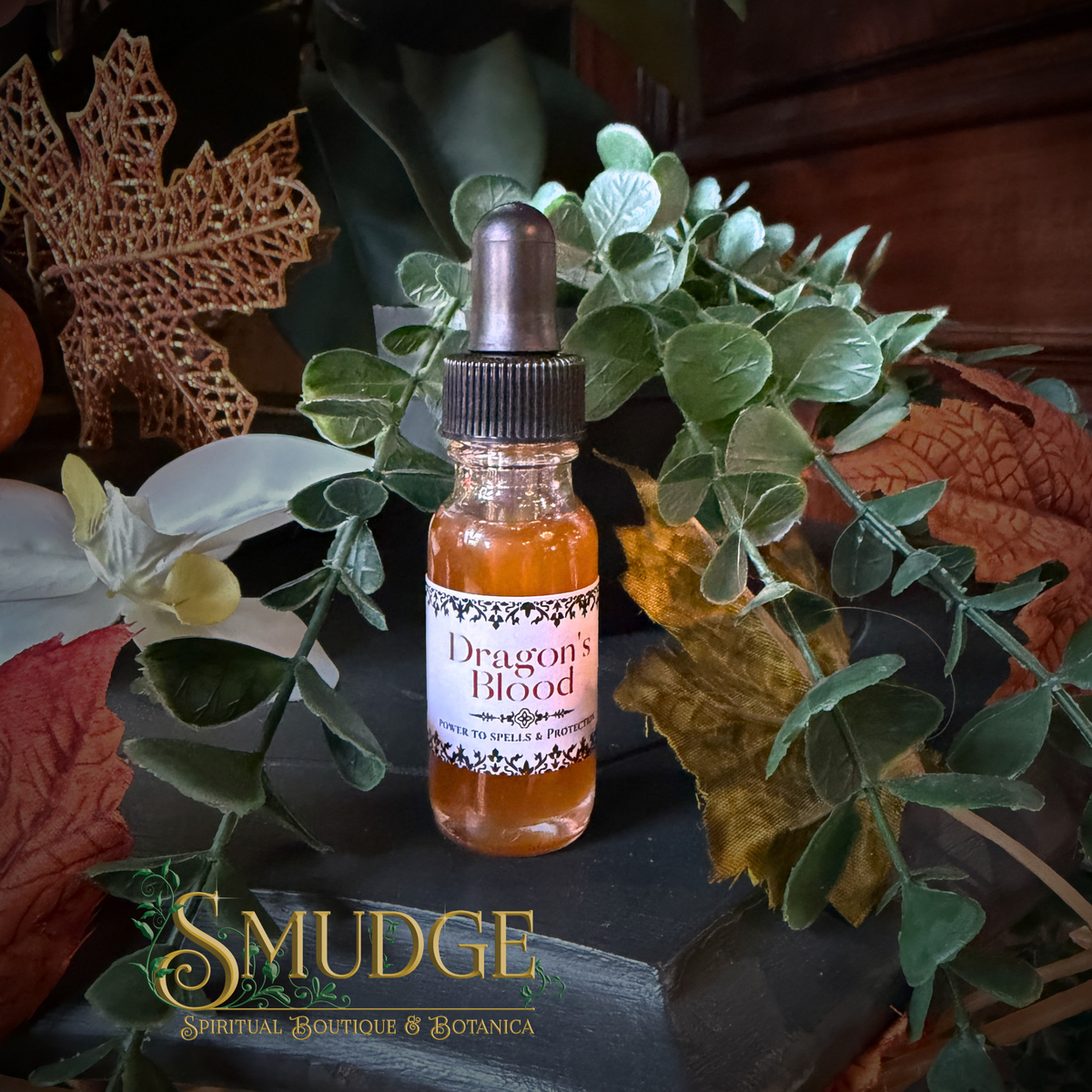 A small glass dropper bottle labeled 'Dragon's Blood' by Smudge Spiritual Boutique & Botanica, filled with amber liquid, styled among green leaves, dried leaves, and a white orchid on a dark surface.