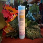 A white pillar candle with a pastel pink label reading 'Dreams' and 'Relax Energy Chakra Candle' stands upright in a styled arrangement with large green leaves, orange flowers, and a red surface. The brand name 'SMUDGE' appears in the lower right corner.