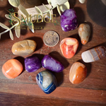 A group of polished tumbled crystals and stones in purple, orange, blue, white, and brown tones arranged on a wooden surface, with a coin and small leaves used for scale and styling. The brand name 'SMUDGE' is visible as an overlay.