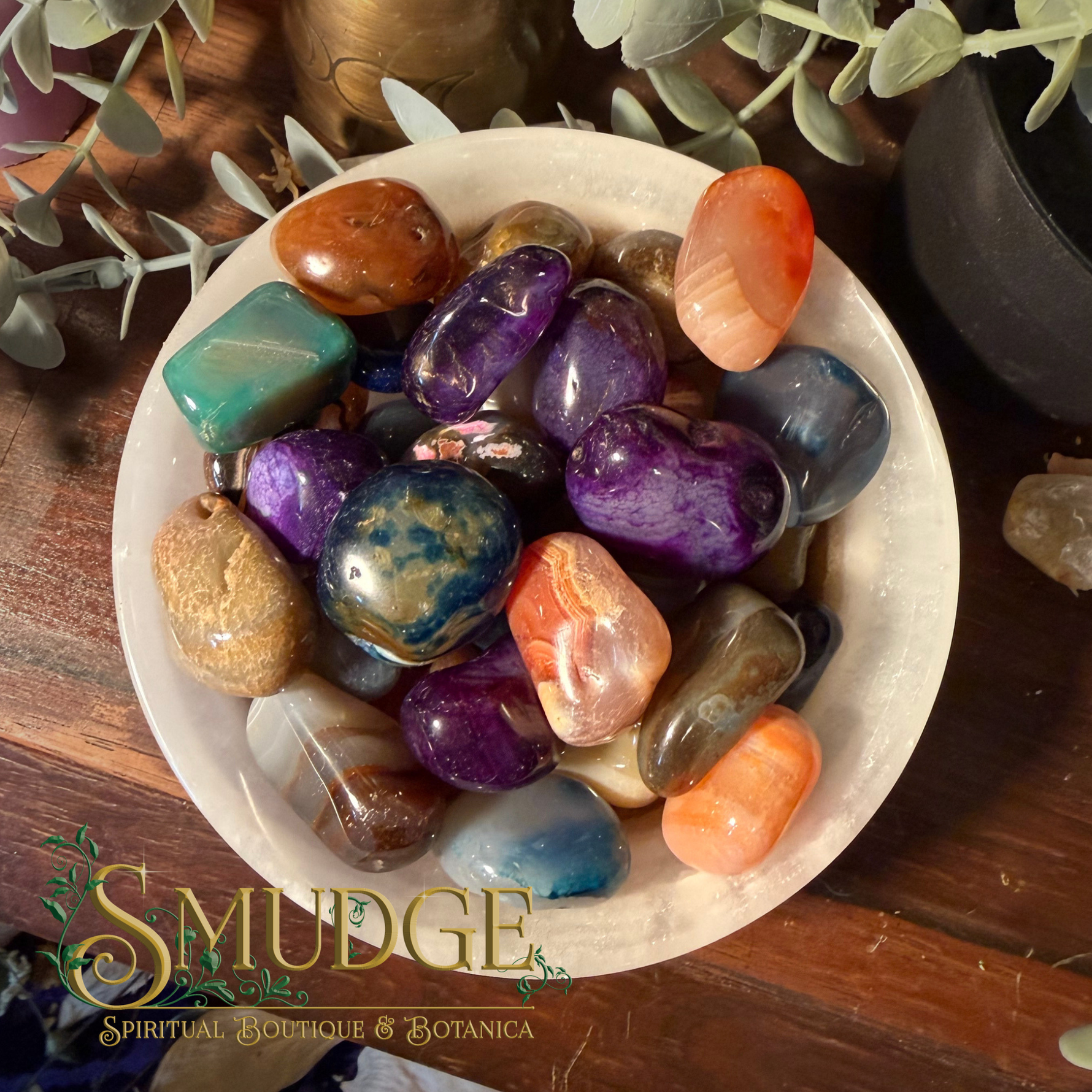A collection of polished tumbled crystals in various colors, including purple, blue, orange, green, and brown, arranged on a white ceramic plate. The plate is set on a dark wooden surface with decorative eucalyptus leaves and a brass candle holder in the background.