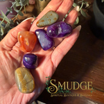 A hand holds five polished tumbled gemstones in various colors including purple, orange, green, and yellow-brown, with a 'SMUDGE Spiritual Boutique & Botanica' logo overlay in the bottom right corner.
