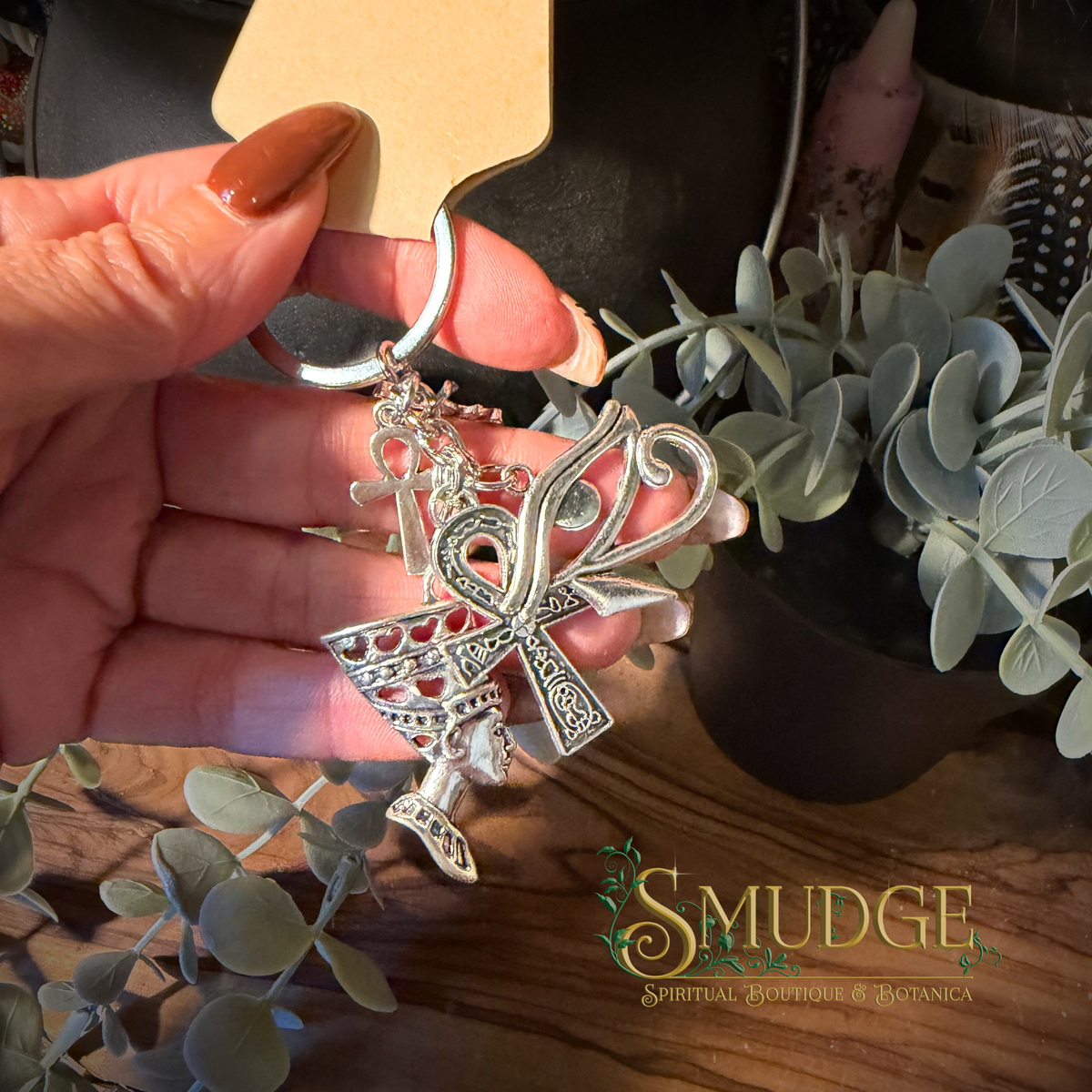 A hand holding a silver-toned keychain featuring an ankh and crossed tools, with a wooden display card attached. The background shows a wooden surface with eucalyptus leaves and a black container, and a 'Smudge Spiritual Boutique & Botanica' logo is visible in the bottom right corner.