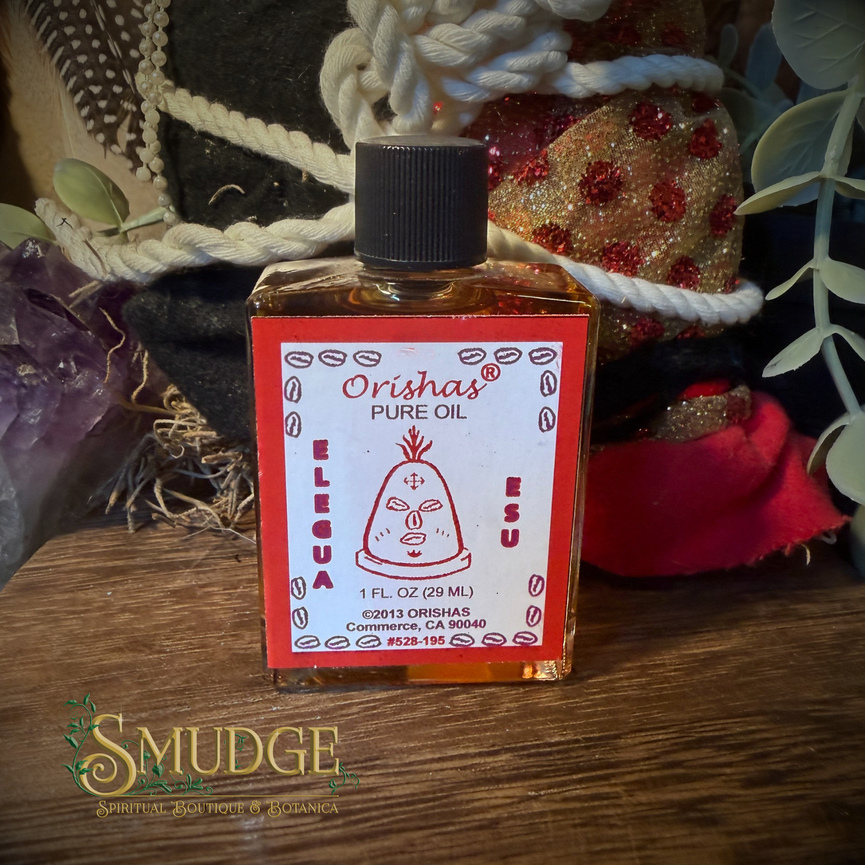 A bottle of Orisha Pure Oil (Elubua) by Smudge Spiritual Wax House & Paraffin Co., placed on a wooden surface with a decorative background of ropes, fabric, and greenery.