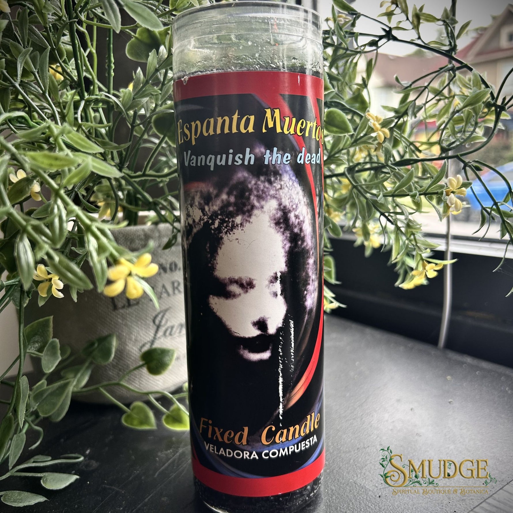 Espanta Muertos Candle with a label featuring a black and white face design, surrounded by green plants.