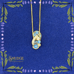 A gold-tone pendant necklace featuring blue and white evil eye motifs, displayed on a textured dark blue background with decorative floral borders and the brand name 'SMUDGE' in the lower left.