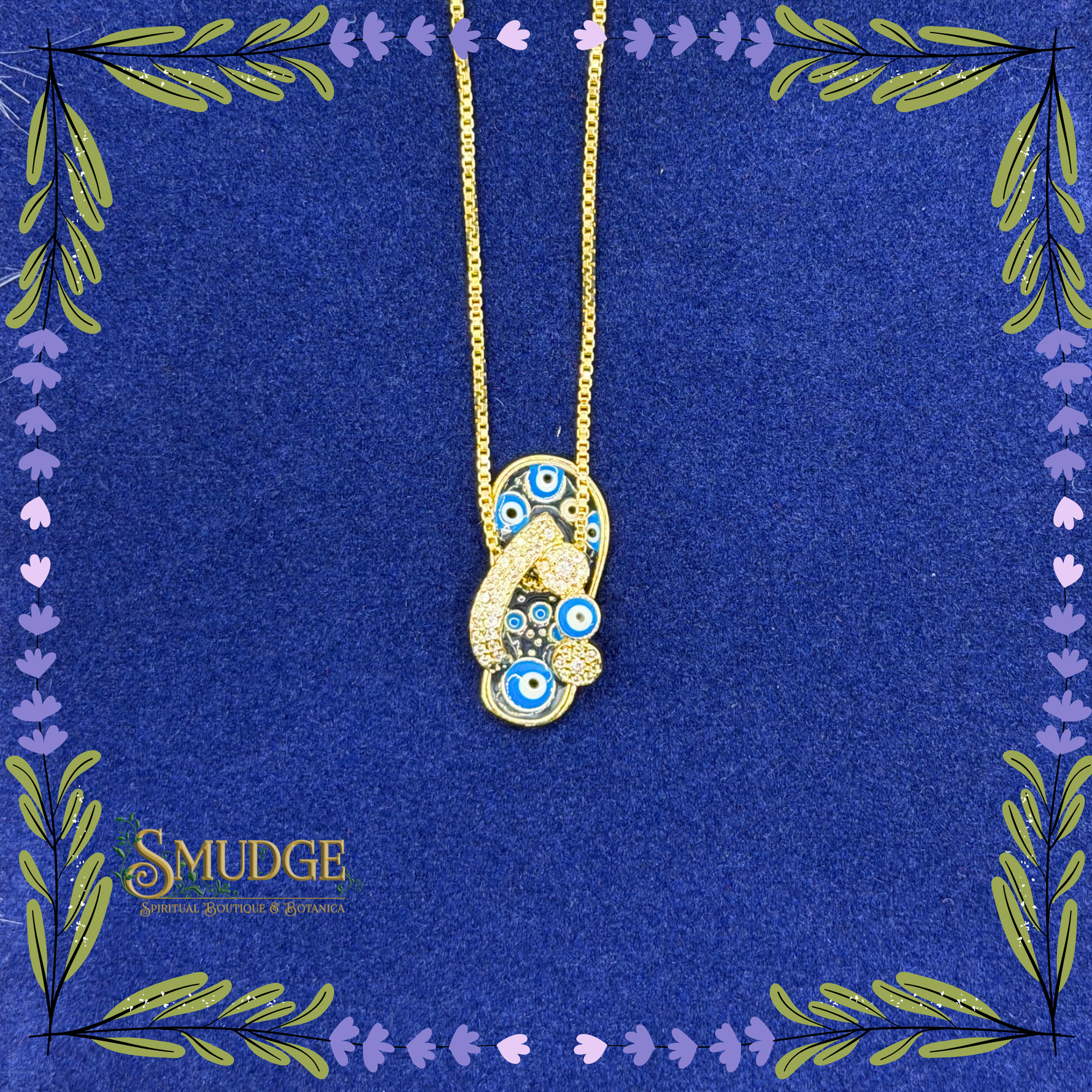 A gold-tone pendant necklace featuring blue and white evil eye motifs, displayed on a textured dark blue background with decorative floral borders and the brand name 'SMUDGE' in the lower left.
