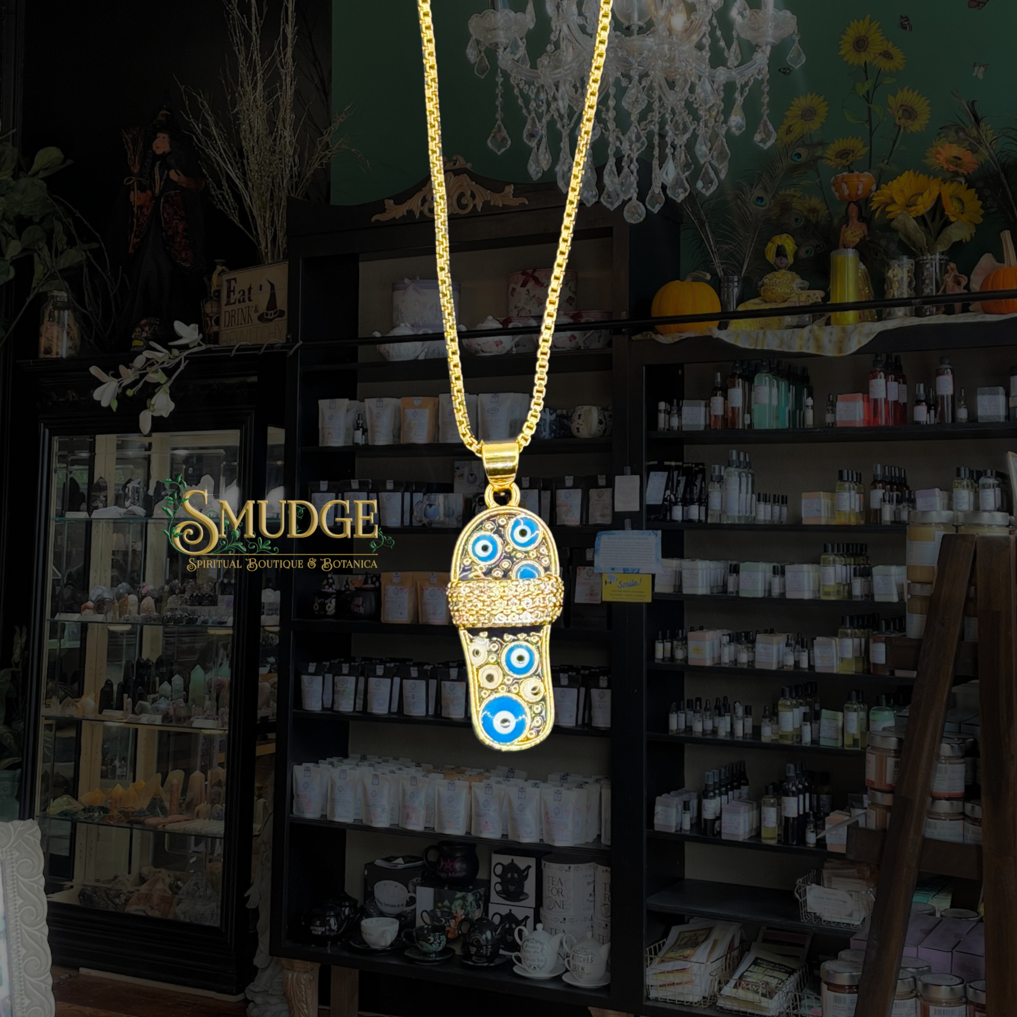 A gold-toned necklace with a flip-flop-shaped pendant featuring blue and white evil eye motifs, hanging in front of a dark shop interior with shelves and a visible MUDGE logo.
