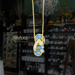 A gold chain necklace with a large, ornate gold-toned pendant featuring blue and white evil eye motifs, hanging in front of a dark boutique interior with shelves of products and a visible 'SMUDGE' sign.