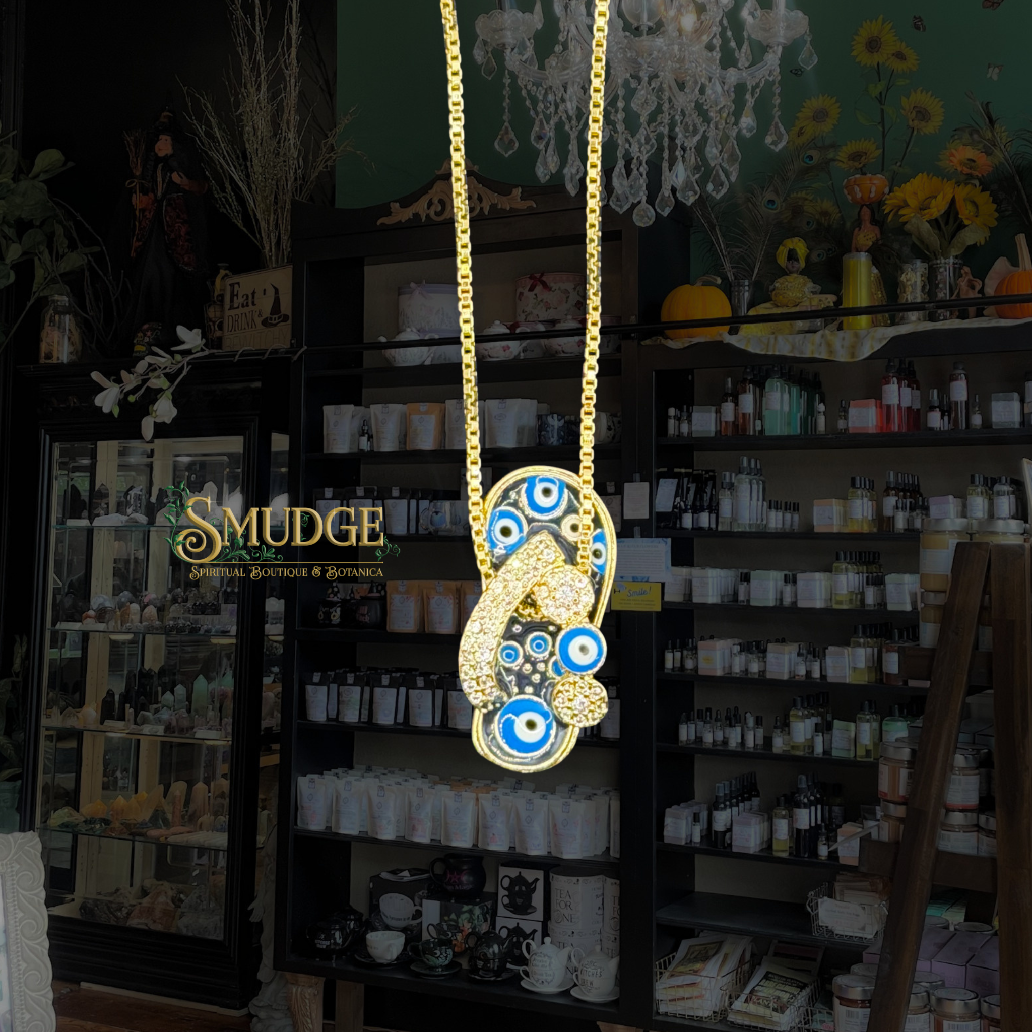 A gold chain necklace with a large, ornate gold-toned pendant featuring blue and white evil eye motifs, hanging in front of a dark boutique interior with shelves of products and a visible 'SMUDGE' sign.