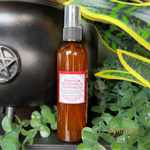 An amber glass spray bottle labeled 'Exorcism, Purification, Uncrossing' with a pink and white label, styled among green plants and a black cauldron on a dark surface.