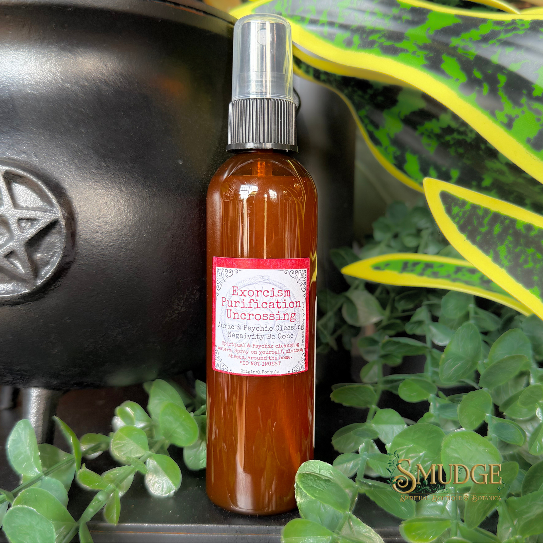 An amber glass spray bottle labeled 'Exorcism, Purification, Uncrossing' with a pink and white label, styled among green plants and a black cauldron on a dark surface.