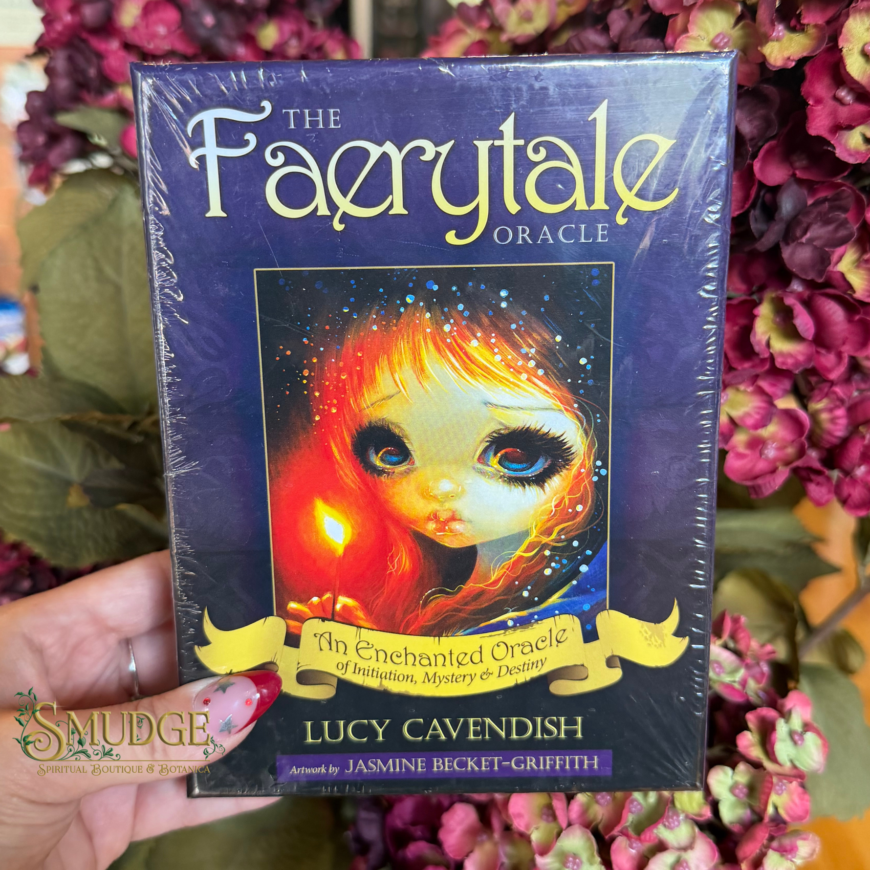 A hand holds a shrink-wrapped copy of 'The Faerytale Oracle' card deck by Lucy Cavendish, set against a background of pink flowers and green leaves. The box features a dark purple background with a central illustration of a fantasy doll with fiery red hair and large blue eyes.