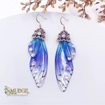 A pair of dangling feather-style earrings with blue, purple, and white wing designs, accented with rhinestones and topped with ornate silver filigree, displayed on a white surface with dried flowers in the background. The brand name 'SMUDGE' is visible in the bottom-left corner.