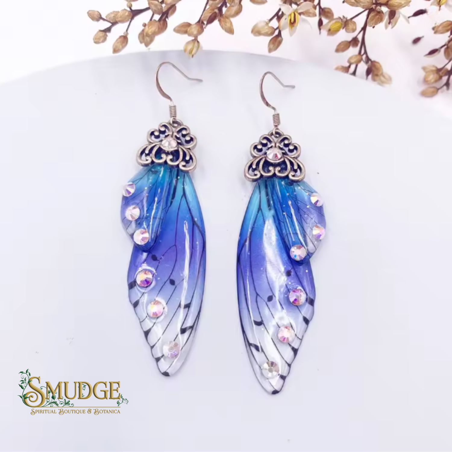 A pair of dangling feather-style earrings with blue, purple, and white wing designs, accented with rhinestones and topped with ornate silver filigree, displayed on a white surface with dried flowers in the background. The brand name 'SMUDGE' is visible in the bottom-left corner.