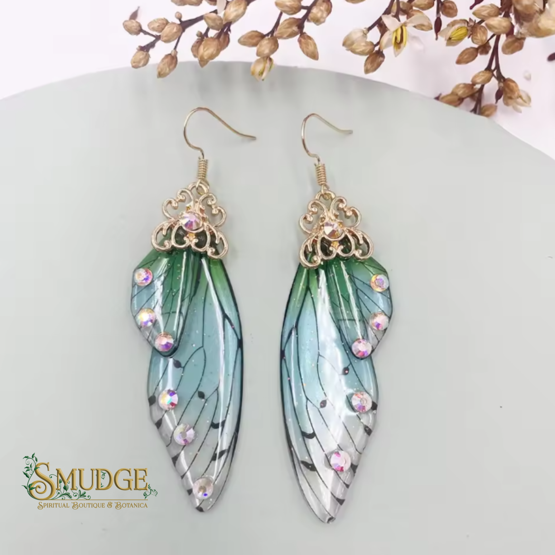 A pair of dangling wing-shaped earrings displayed on a pale gray surface with decorative dried leaves at the top. The earrings have gold-tone filigree tops, iridescent rhinestones, and translucent green, blue, black, and white wing designs.