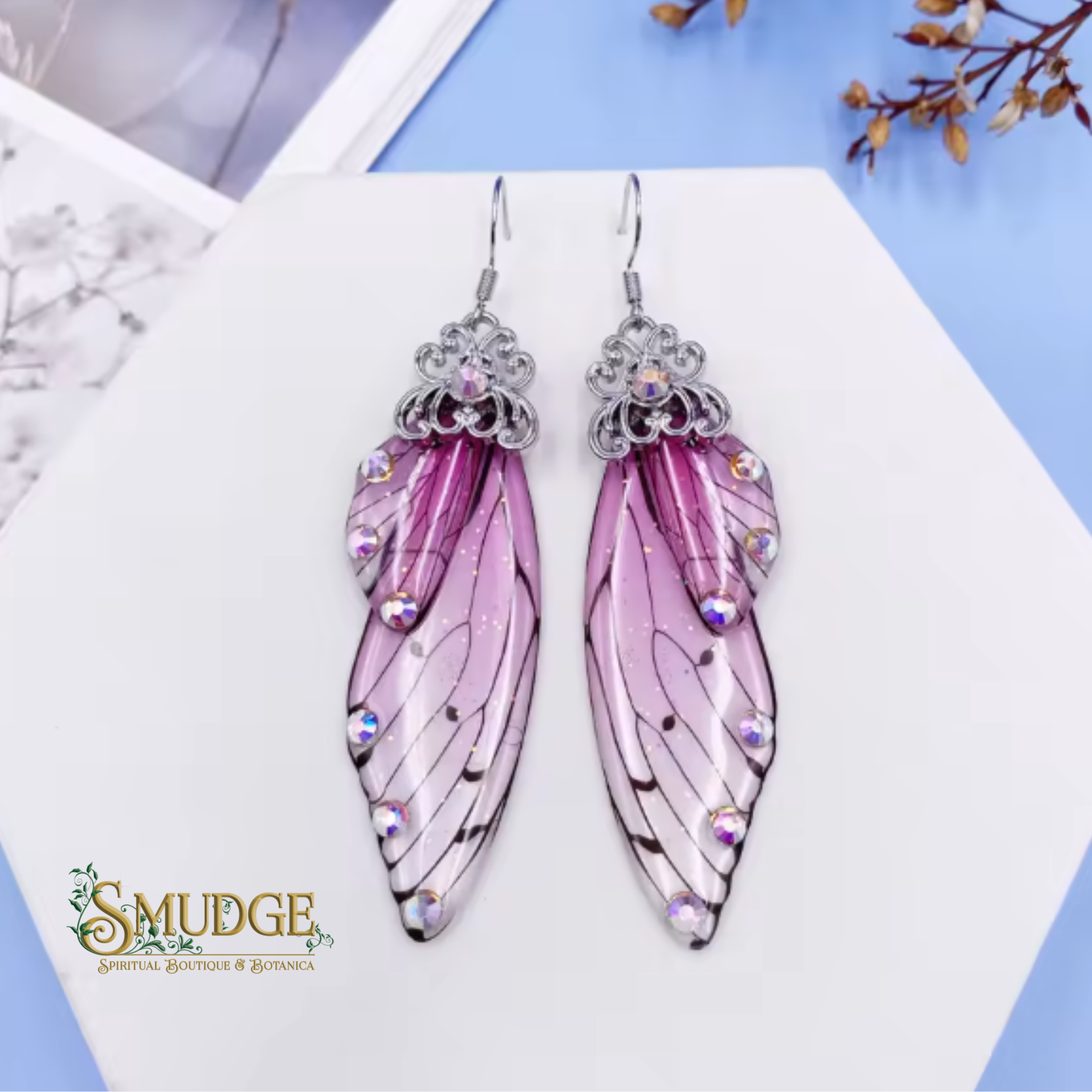 A pair of dangling wing-shaped earrings with pink and purple translucent wings, silver filigree tops, and iridescent rhinestones, displayed on a white card with a blue background and the Smudge brand logo visible.