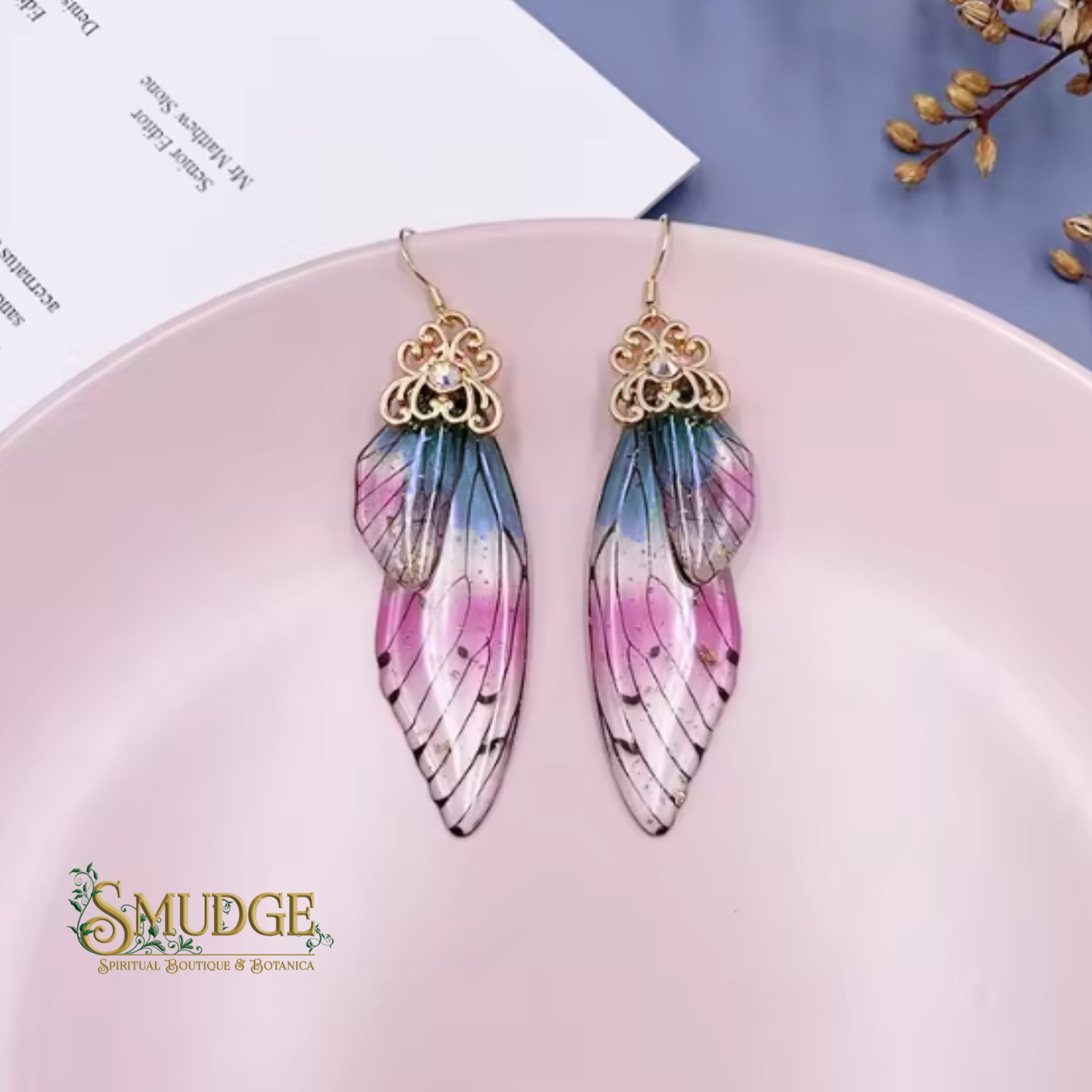 A pair of dangling wing earrings with gold-tone filigree tops and wings featuring a pink, blue, and white gradient, displayed on a light pink plate.