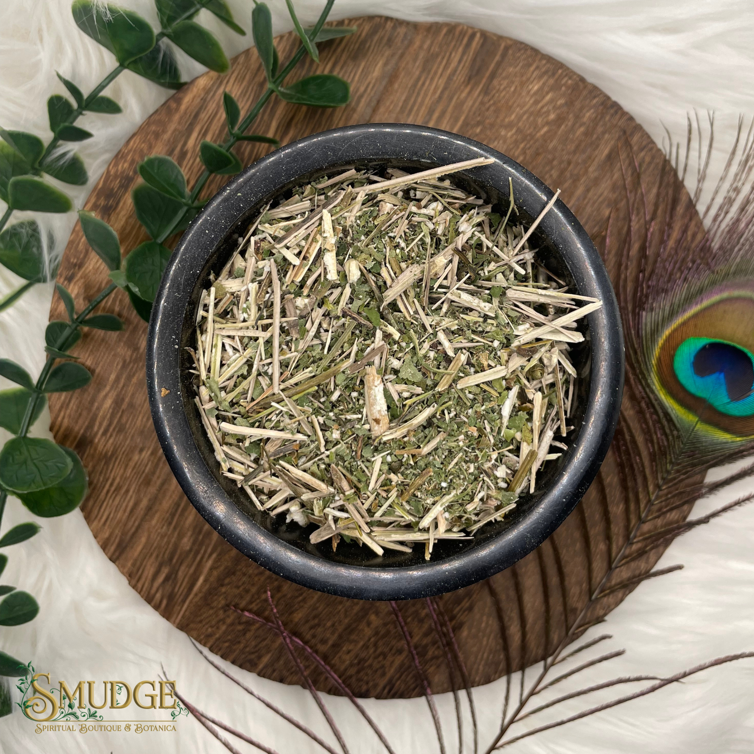 Dried herbs in a black bowl on a wooden tray with peacock feather and green leaves.