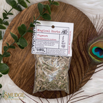 Bag of dried herbs labeled 'Magical Herbs' on a wooden board with peacock feather and green leaves.