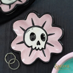 A pink flower-shaped ceramic dish with a white skull and black outlines on a dark textured surface. A small silver key ring is placed beside it, and part of another matching dish is visible in the top-left corner.