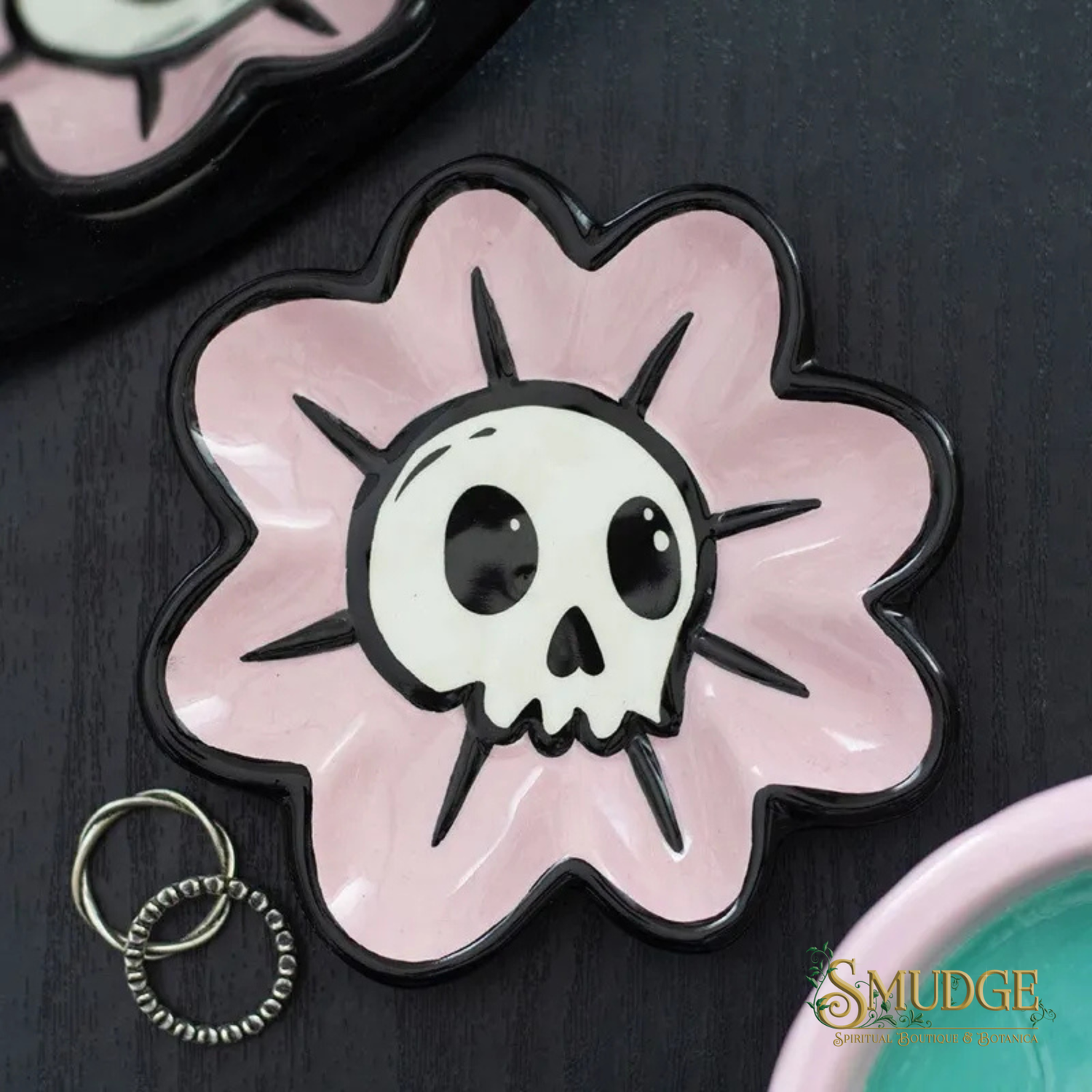 A pink flower-shaped ceramic dish with a white skull and black outlines on a dark textured surface. A small silver key ring is placed beside it, and part of another matching dish is visible in the top-left corner.
