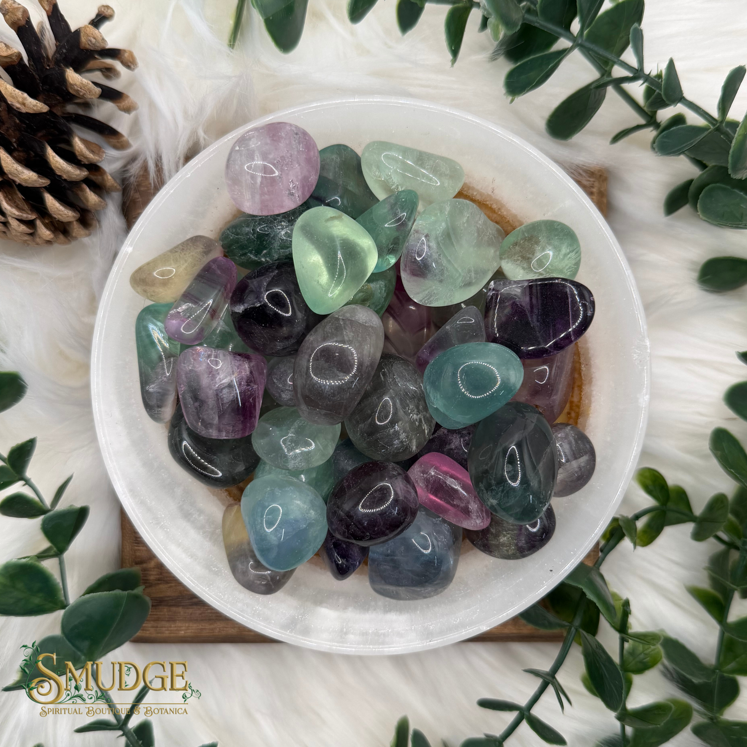 A white bowl filled with polished tumbled gemstones in assorted pastel and deep colors, arranged on a wooden board with green leaves and a pinecone around it. The SMUDGE Gemstone Collection logo is visible in the lower-left corner.