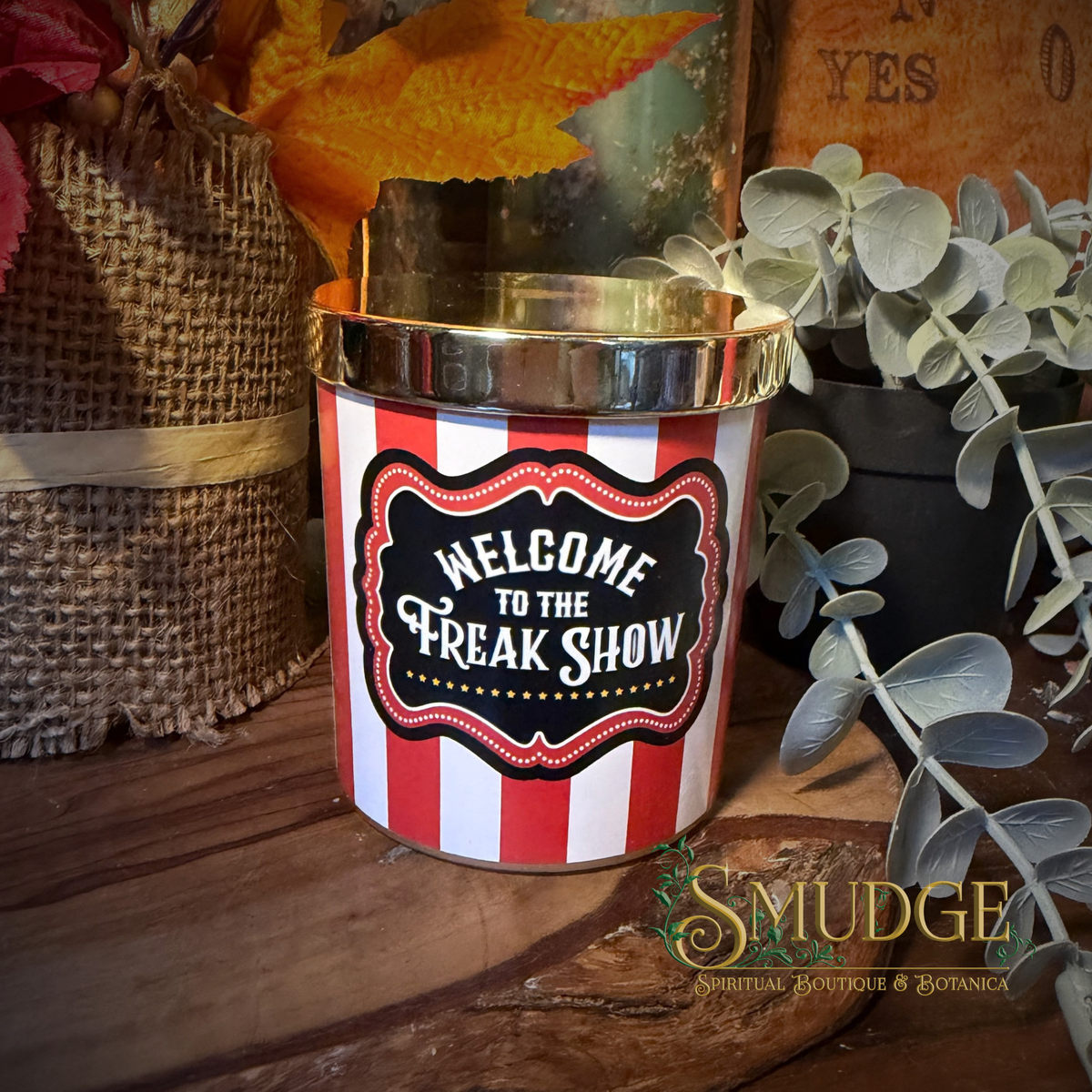 A cylindrical tin candle or scented vessel with a red and white striped body and a shiny gold-toned rim, featuring a front label that reads 'WELCOME TO THE FREAK SHOW.' It is styled on a wooden surface with burlap, dried leaves, greenery, and rustic decor in the background.