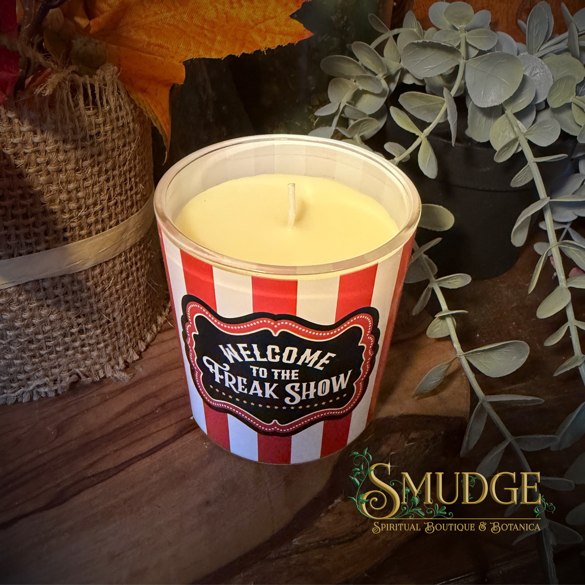A novelty jar candle with a red and white striped design and a black label reading 'Welcome to the Freak Show' sits on a rustic wooden surface. The candle is styled with a burlap bag, autumn leaves, and green eucalyptus branches.