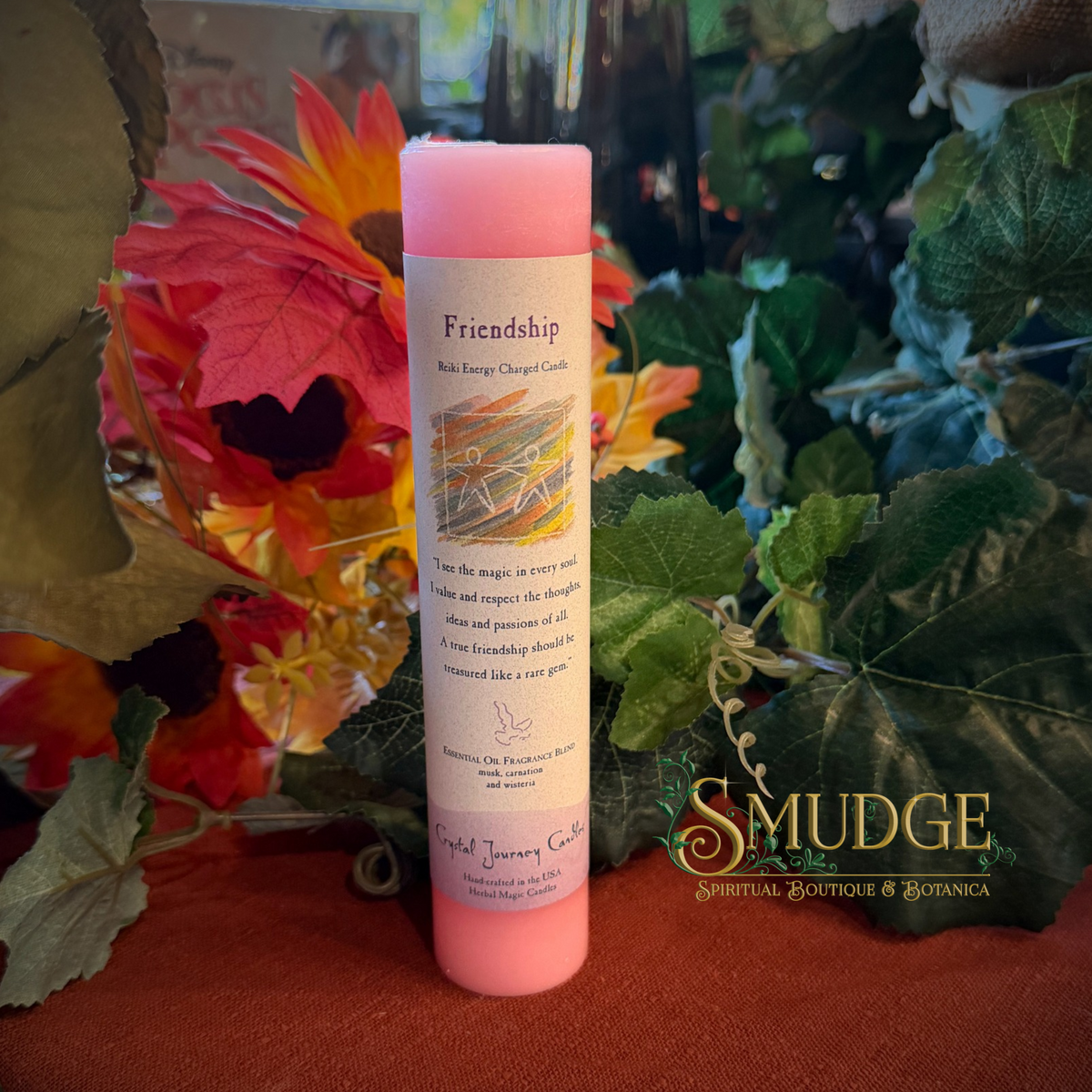 A pink bath energy charge candle labeled 'Friendship' stands upright on a red surface surrounded by autumn leaves and green foliage. The candle wrapper has a white label with text and a small illustration, and a watermark reading 'SMUDGE' appears on the right side.