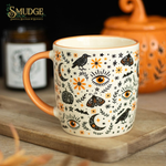 A ceramic mug with a white base decorated with black and orange Halloween-themed illustrations including eyes, crows, moons, and moths, resting on a wooden cutting board with a blurred background.