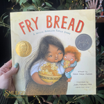 Book titled 'Fry Bread' held by a hand with green nail polish, surrounded by greenery.