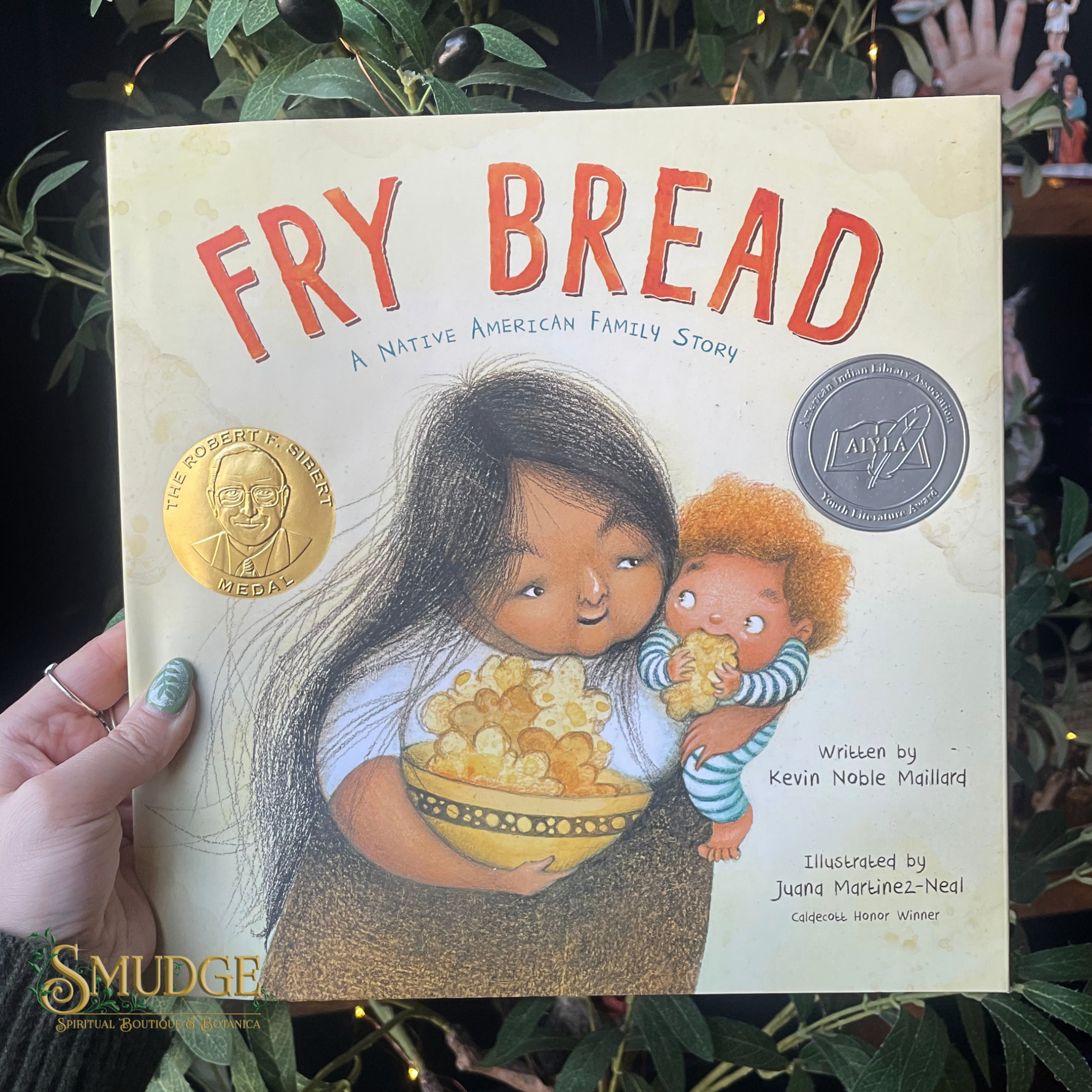 Book titled 'Fry Bread' held by a hand with green nail polish, surrounded by greenery.