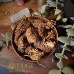 A brown ceramic bowl filled with dried galangal roots and botanical pieces, styled on a wooden surface with eucalyptus leaves, crystals, and a white stone. The brand name 'SMUDGE' is visible in the bottom-left corner.