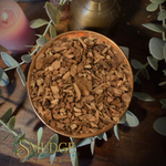 A top-down view of a copper bowl filled with galangal root chips or wood shavings, surrounded by green eucalyptus leaves on a dark wooden surface. A pink candle is visible in the upper left, and a brass container is in the background.