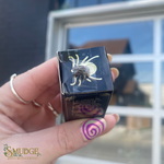 Hand holding a small black box with a white design, labeled 'Smudge', against a blurred outdoor background.