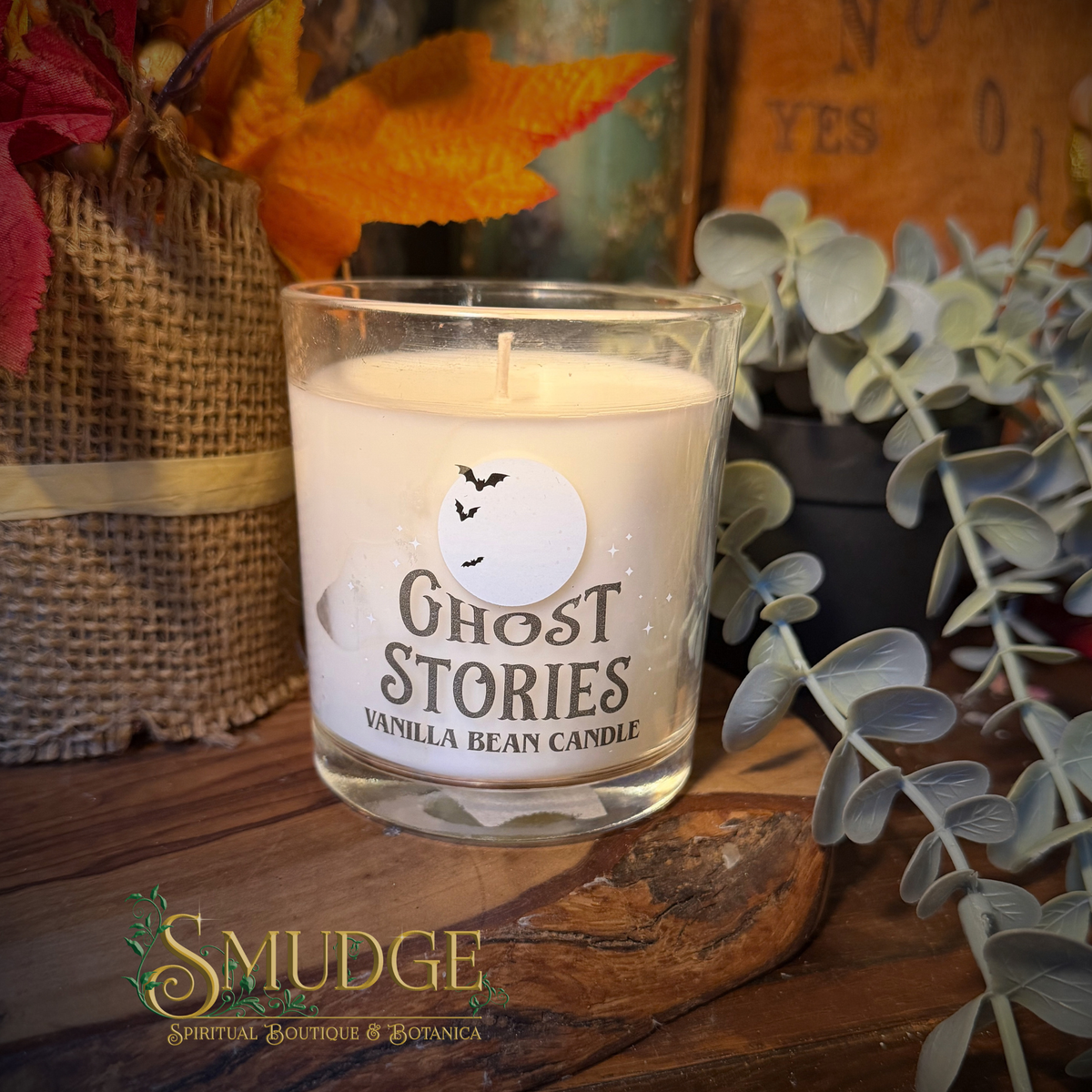 A glass jar candle labeled 'Ghost Stories Vanilla Bean Candle' sits on a rustic wooden surface, surrounded by eucalyptus leaves, burlap, and autumn leaves. The candle wax is off-white, and the label features a white moon graphic with black bats.
