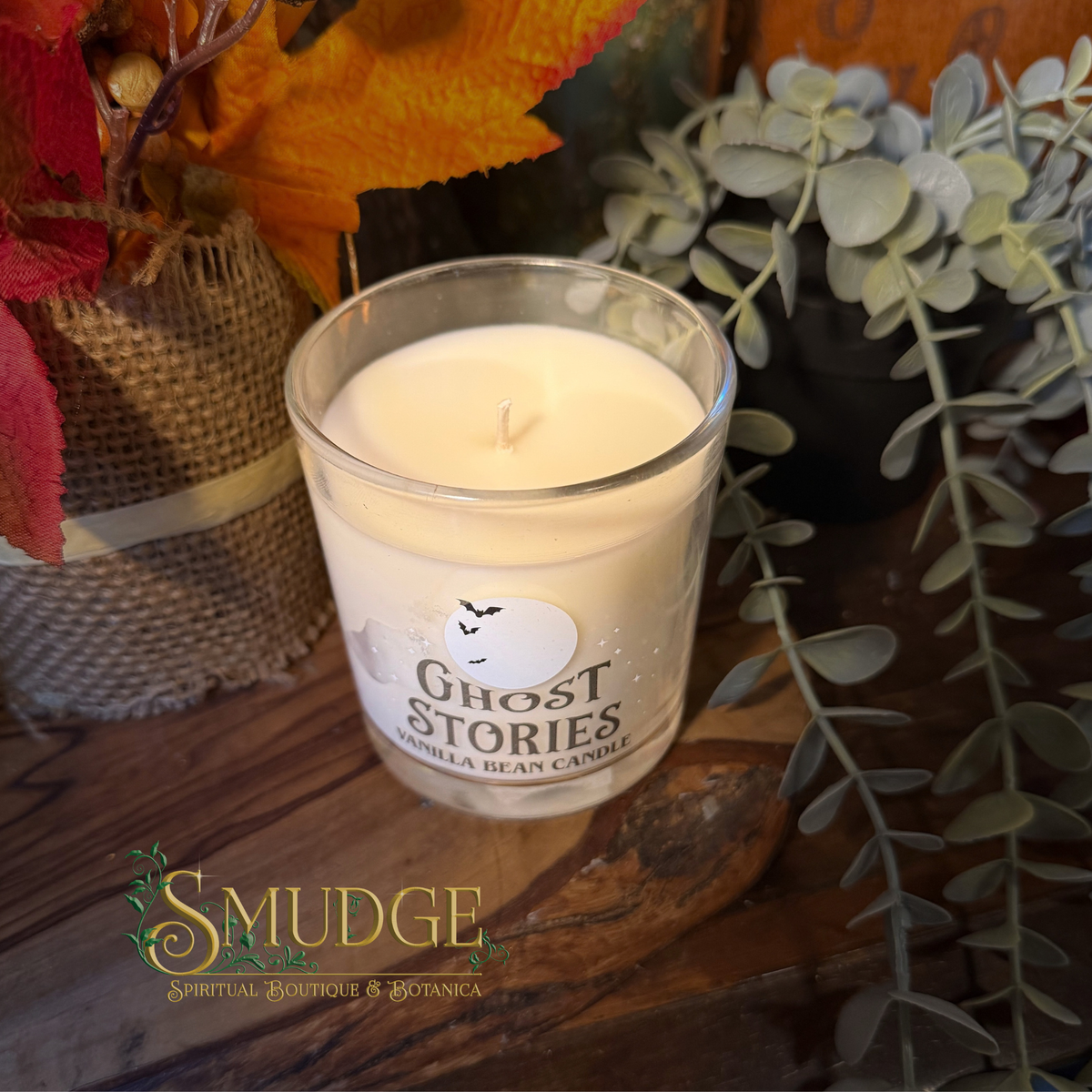 A glass jar candle labeled 'Ghost Stories' sits on a rustic wooden surface surrounded by autumn leaves, burlap, and green foliage. The candle wax is off-white, and the label features a ghost illustration.