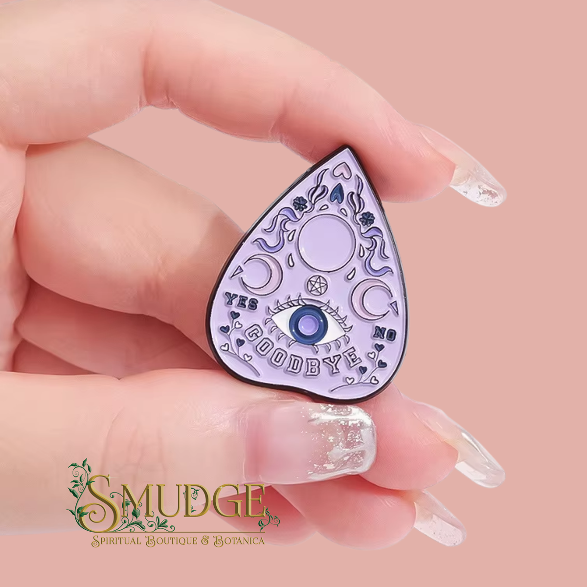 A hand with long clear acrylic nails holds a teardrop-shaped enamel pin featuring a moon and star design with the text 'TYS GOODBYE' and a blue eye motif. The background is a solid dusty pink, and a 'SMUDGE Spiritual Boutique & Botanica' logo is visible in the bottom-left corner.
