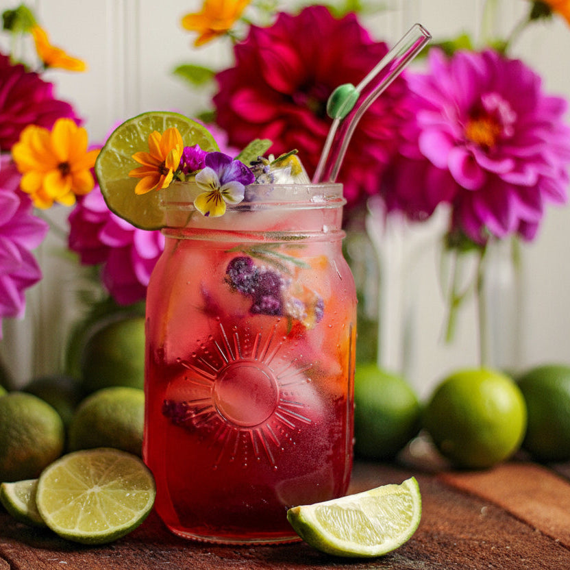 A mason jar filled with a bright pink-red iced drink, garnished with a lime wheel, edible flowers, and a glass straw, set on a wooden table with limes and colorful flowers in the background.