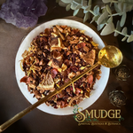 A top-down view of a white ceramic bowl filled with a colorful dried botanical blend, with a gold spoon resting inside. The scene is styled with a purple crystal cluster, green leaves, and decorative metal ornaments on a dark wooden surface.