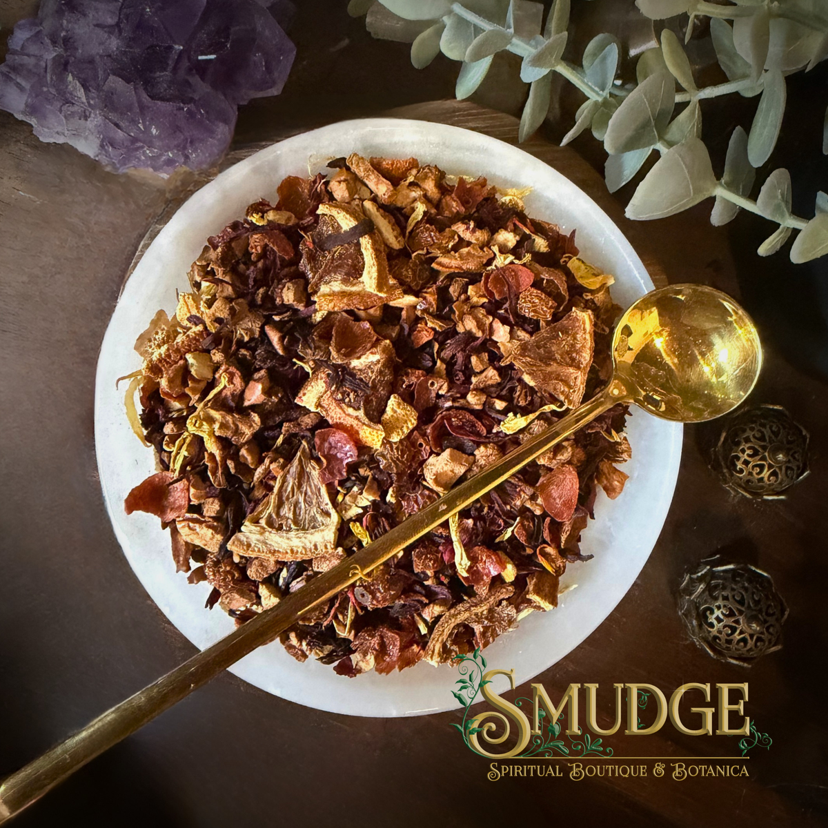 A top-down view of a white ceramic bowl filled with a colorful dried botanical blend, with a gold spoon resting inside. The scene is styled with a purple crystal cluster, green leaves, and decorative metal ornaments on a dark wooden surface.