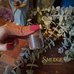 Hand holding a small glass container with herbs, surrounded by eucalyptus leaves and a figurine, on a wooden surface with 'Smudge' branding.